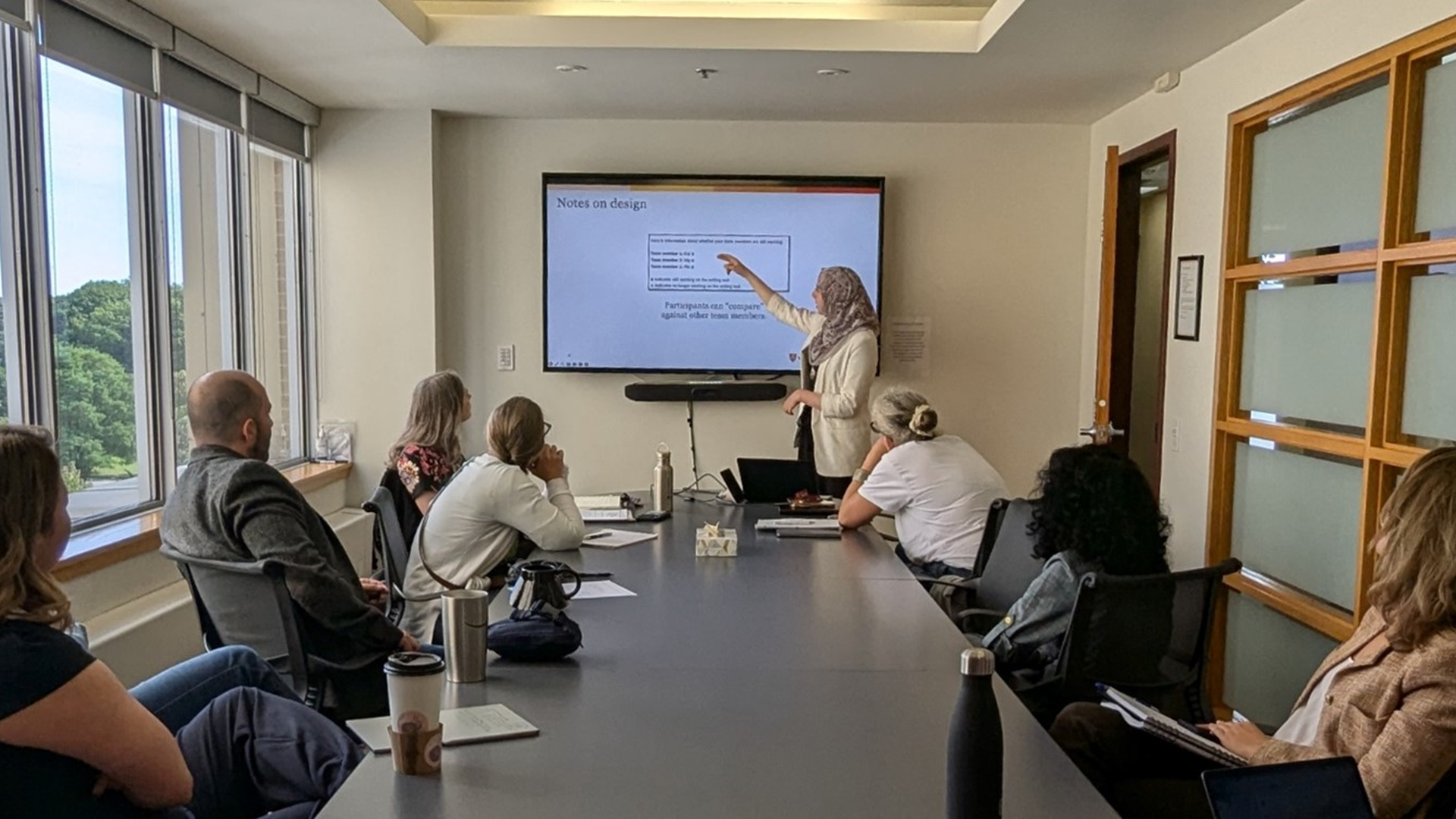 A group of people sitting around a board table. They are paying attention to a person presenting at the front of the room, using a large screen.