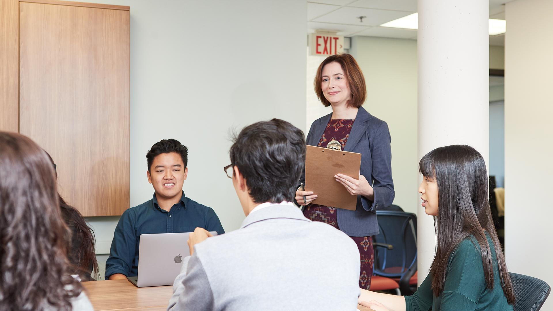 Several people sitting at a table, while a smiling woman stands nearby with a clipboard.