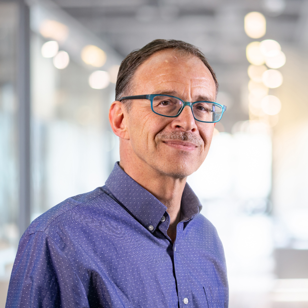 Headshot of Ron Balvers, a smiling man with a moustache, blue glasses, wearing a violet shirt.