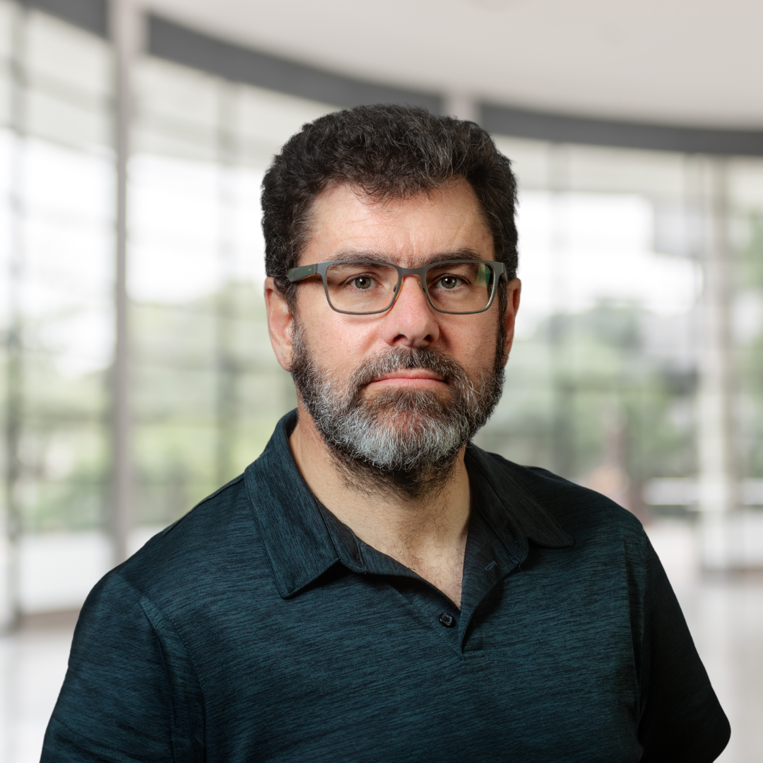 Headshot of John Maheu, a smiling man wearing glasses and a dark teal shirt.