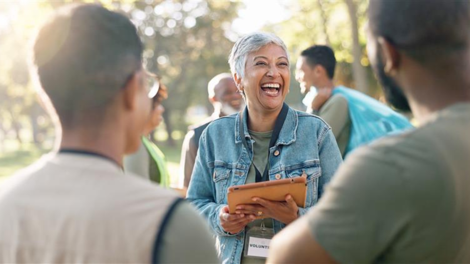 A smiling volunteer holding a tablet laughs while talking with community group members.